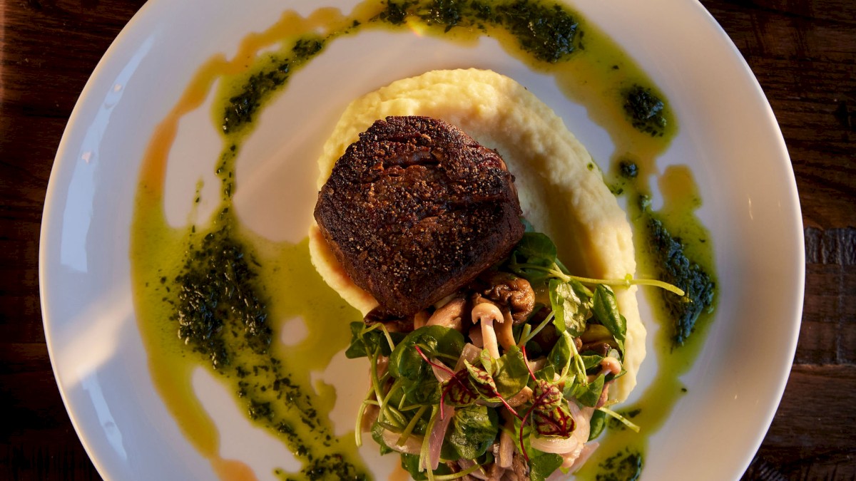 Currents Restaurant at Riverhouse Lodge showcases a beautifully presented dish on a white plate, featuring a steak, mashed potatoes, greens, and sauce, with a glass of white wine nearby.