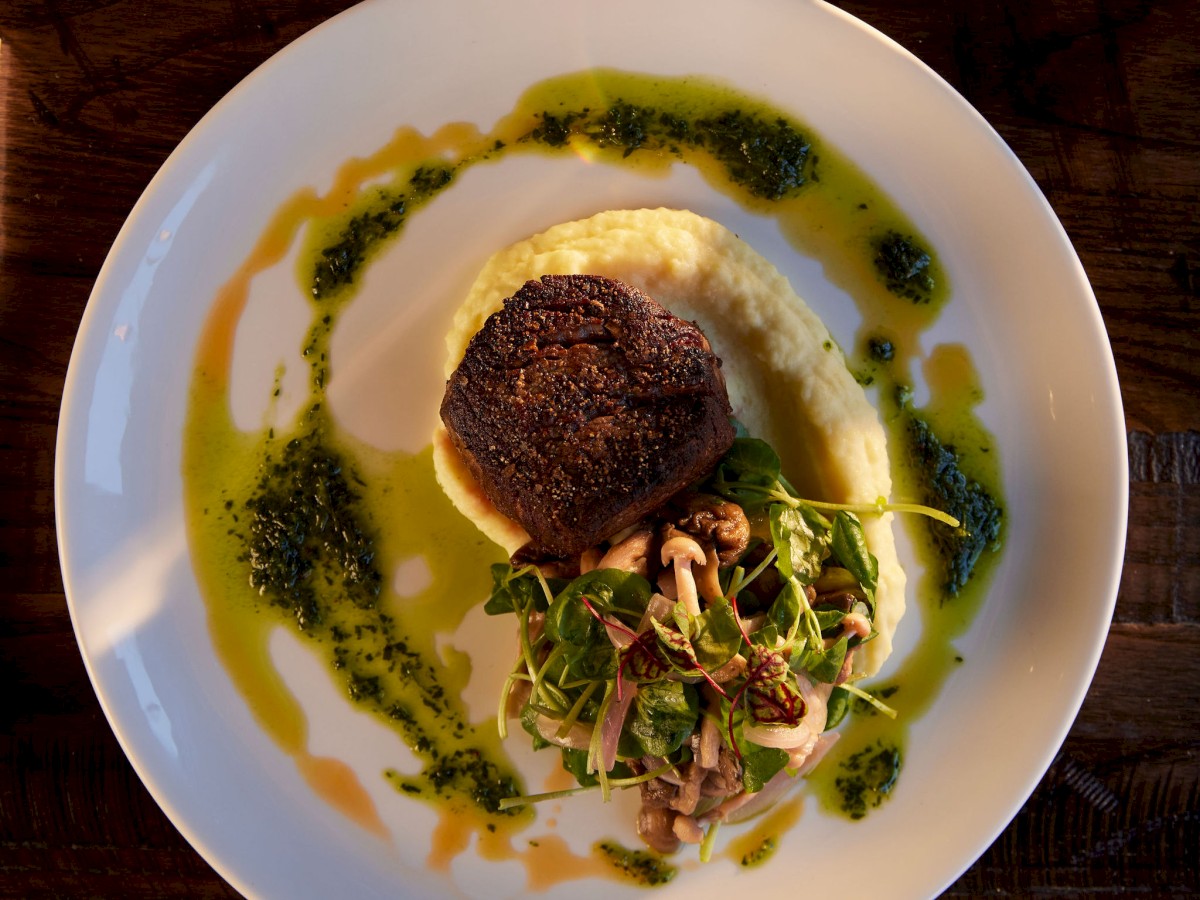 The image shows a beautifully presented dish on a white plate, featuring a steak, mashed potatoes, greens, and sauce, with a glass of white wine nearby.