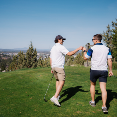 Two people at River's Edge golf course in Bend, Oregon, shaking hands with clubs in hand, surrounded by trees and a distant city view under a clear blue sky.