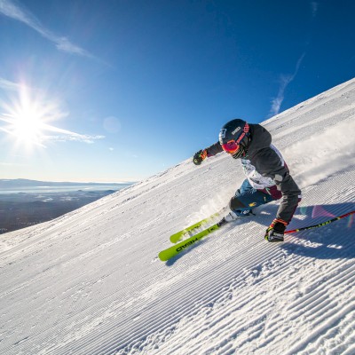 A skier is racing down a snowy slope on a bright, sunny day, kicking up powder behind them under a clear blue sky.