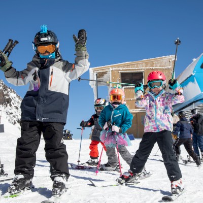 Children and a guide in ski gear stand on a snowy slope, posing with ski poles, near a blue ski lift under a clear blue sky.