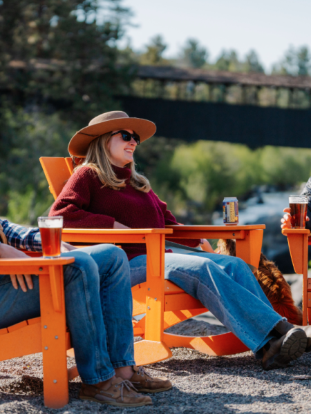 Three people sit outside Riverhouse Lodge in Bend, Oregon in orange chairs, enjoying drinks and chatting by a river with a forest bridge in the background.