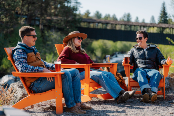Three people sit outside Riverhouse Lodge in Bend, Oregon in orange chairs, enjoying drinks and chatting by a river with a forest bridge in the background.