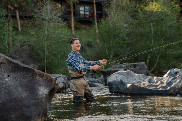 A man with fishing gear stands waist-deep in a river, smiling in a scenic outdoor setting with trees and a house in the background.
