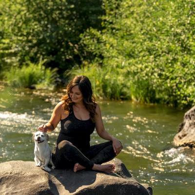 A woman sits on a stone in the Deschutes River that passes through Riverhouse Lodge in Bend, Oregon, smiling while petting a small dog, with greenery around them.