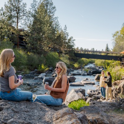 Two women sit on rocks by a river, talking and smiling, with others in the background enjoying a sunny day in nature.