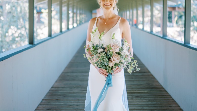 A bride in a white wedding dress stands in a covered walkway, holding a bouquet of flowers tied with a blue ribbon, smiling at the camera.