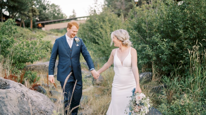 A couple, dressed in wedding attire, walk hand in hand in a lush, green outdoor setting with the bride holding a bouquet and smiling at the groom.