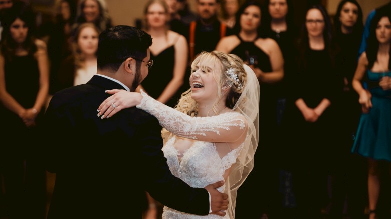 A bride and groom are dancing together, with an audience of guests watching in the background, some of whom are smiling and clapping.