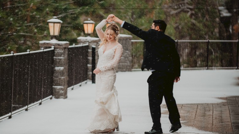 A couple dances together in the snow on a pathway lined with lampposts and surrounded by greenery, with the bride in a white dress and groom in black attire.