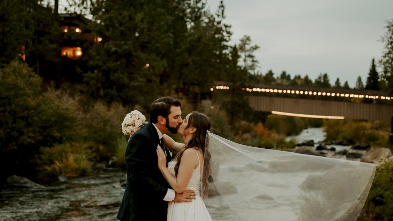 A bride and groom share a kiss outdoors by a river, with trees and a bridge in the background, the bride's veil flowing behind them.