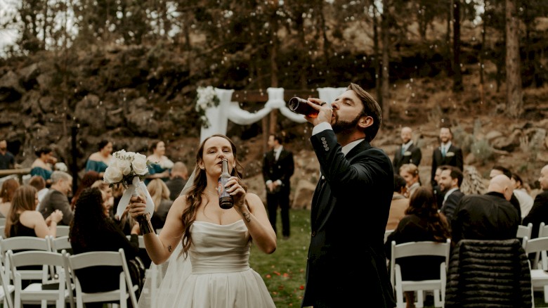A bride and groom are drinking from animal-shaped vessels in an outdoor setting, surrounded by guests seated on white chairs amongst trees.