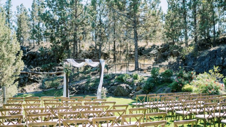 An outdoor wedding setup with rows of wooden chairs centered around an altar adorned with white fabric, in a forested area under clear skies.