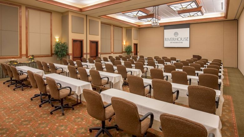 The image shows a conference room with rows of chairs and tables facing a projection screen with the text “Riverhouse on the Deschutes.”