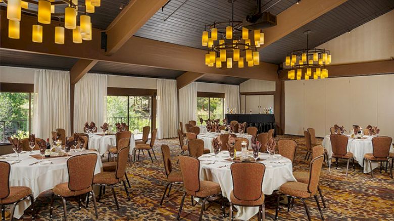 An elegant dining area with round tables, brown chairs, white tablecloths, warm lighting from chandeliers, large windows, and carpeted floor.