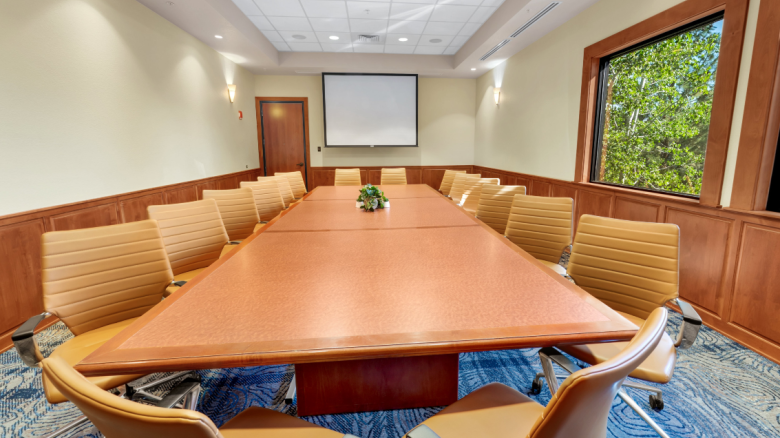 A conference room with a large table, tan chairs, a projector, screen, and window views outside.