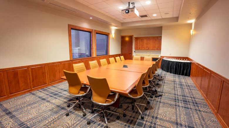 A conference room with a long wooden table, multiple chairs, a projection system, and a side table with refreshments in the corner.