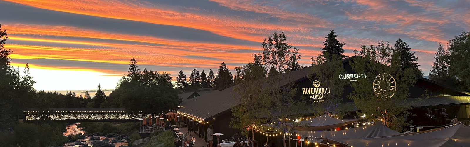 An outdoor dining area beside a river, with people gathered under string lights and a stunning sunset in the background.
