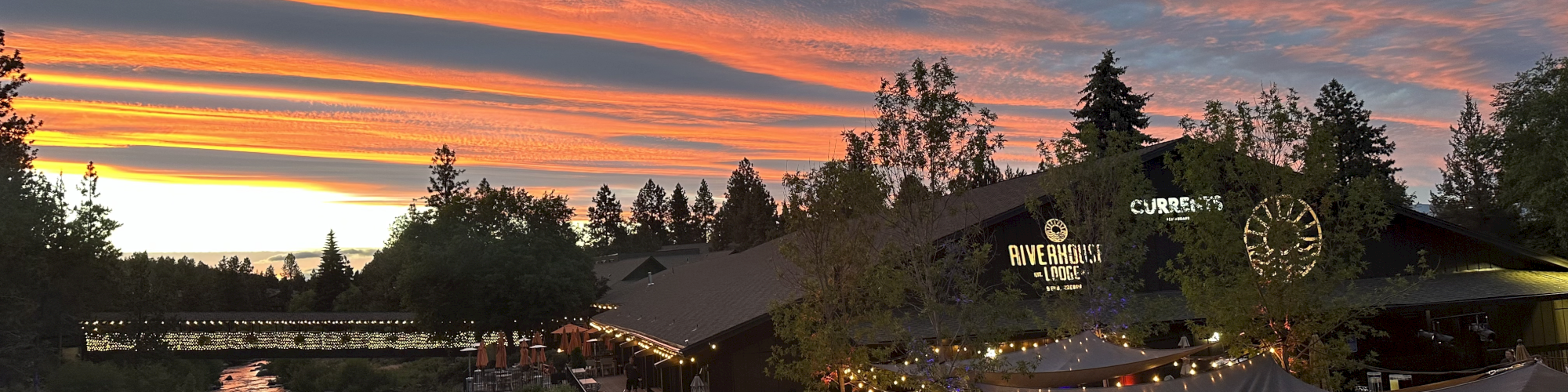 An outdoor dining area beside a river, with people gathered under string lights and a stunning sunset in the background.