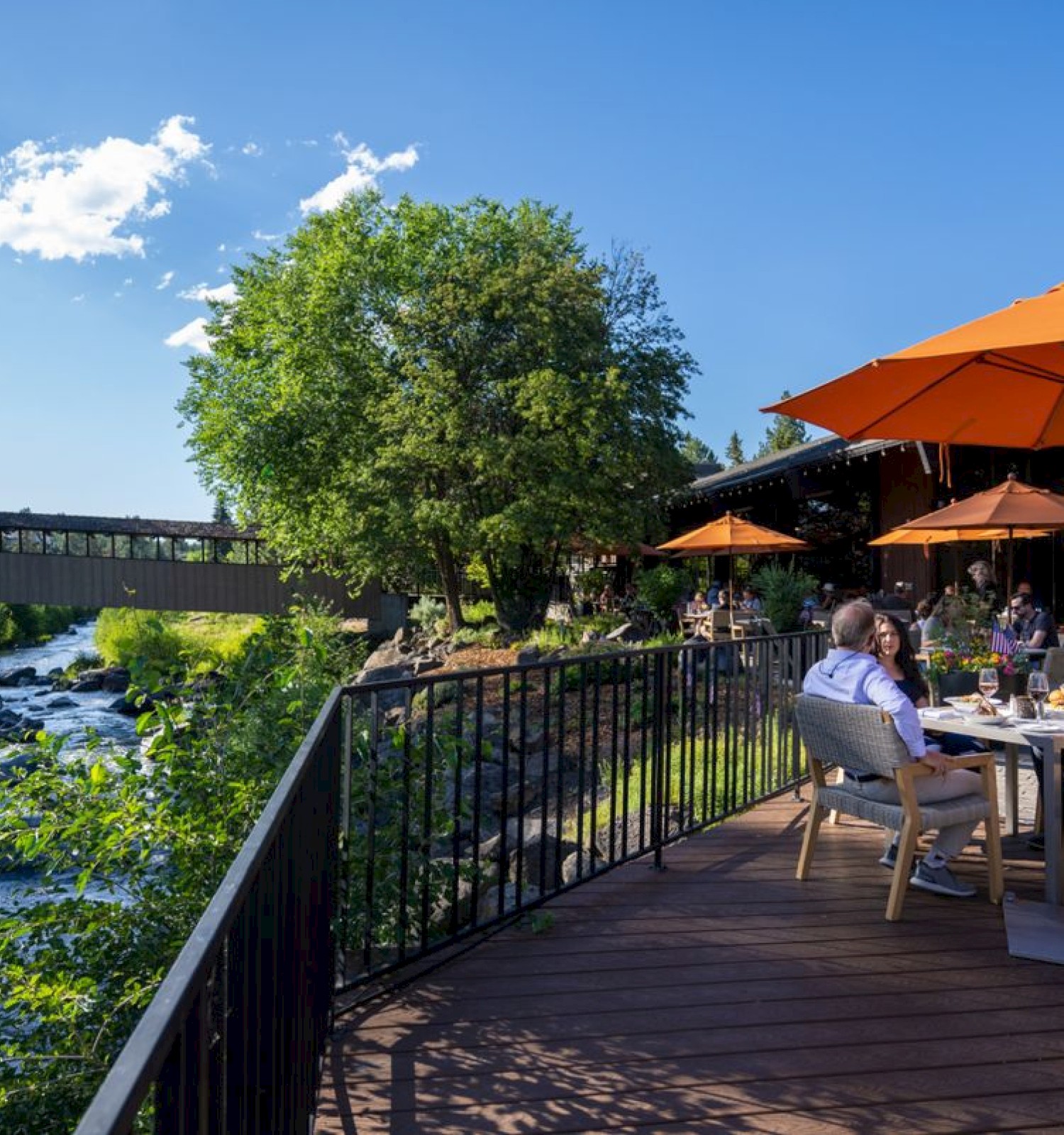 Outdoor dining area by a scenic river with people sitting under orange umbrellas, surrounded by trees and a wooden bridge.