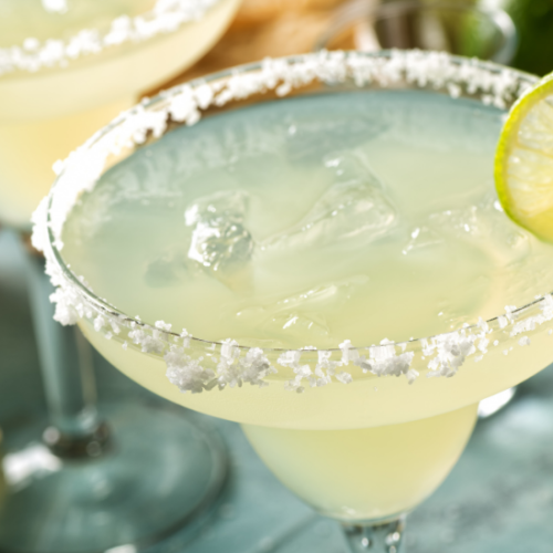 A close-up of a margarita cocktail in a glass with a salted rim and a lime slice garnish, with another similar drink in the background.