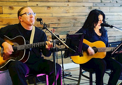 Two musicians are performing with guitars, seated in a cozy setting. A sign in the front reads 