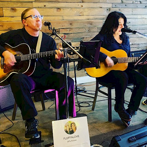 Two musicians are performing with guitars, seated in a cozy setting. A sign in the front reads 