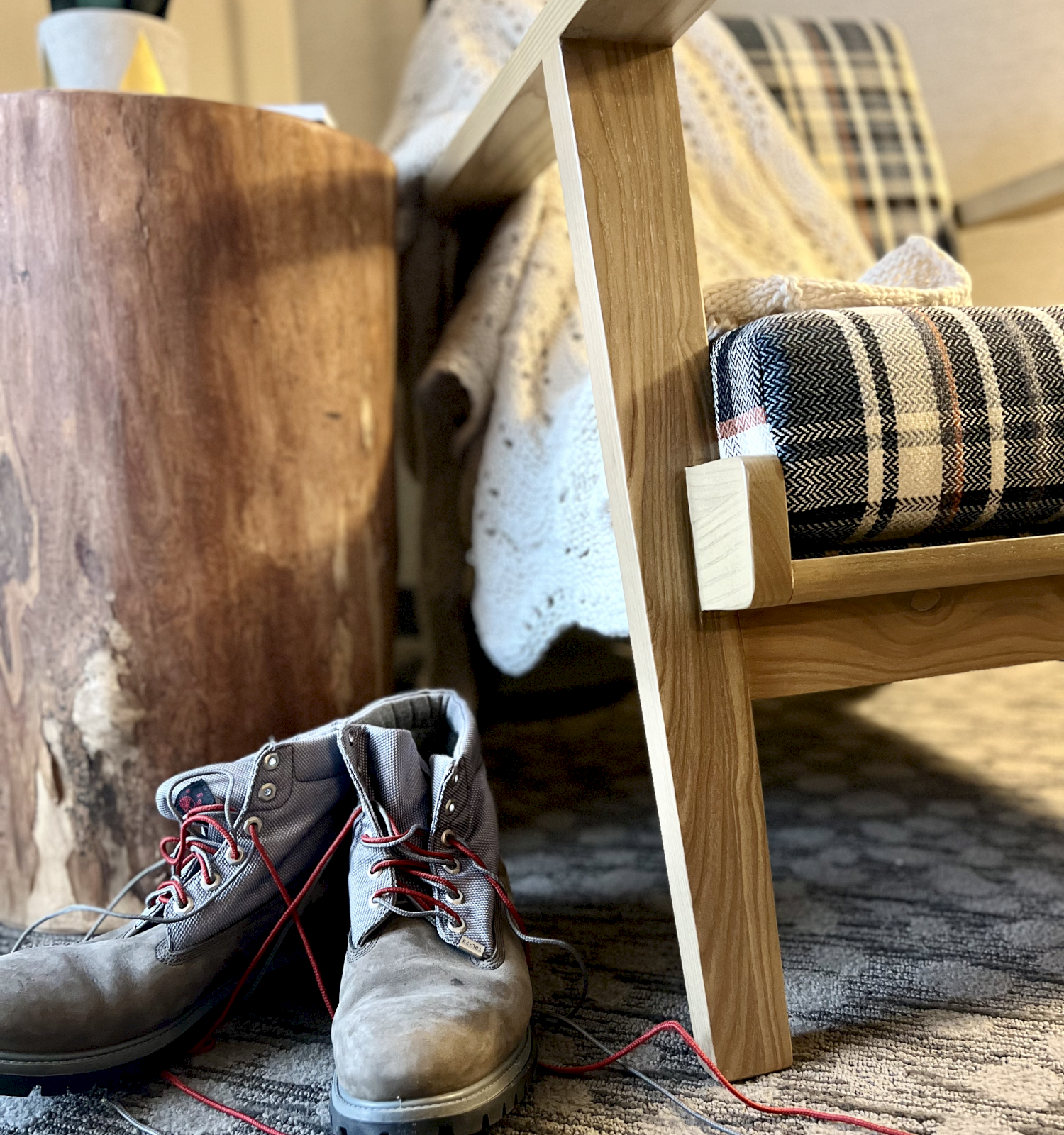 A cozy scene with a wooden chair, plaid blanket, tree stump sidetable, and a pair of boots with red laces on a patterned carpet.