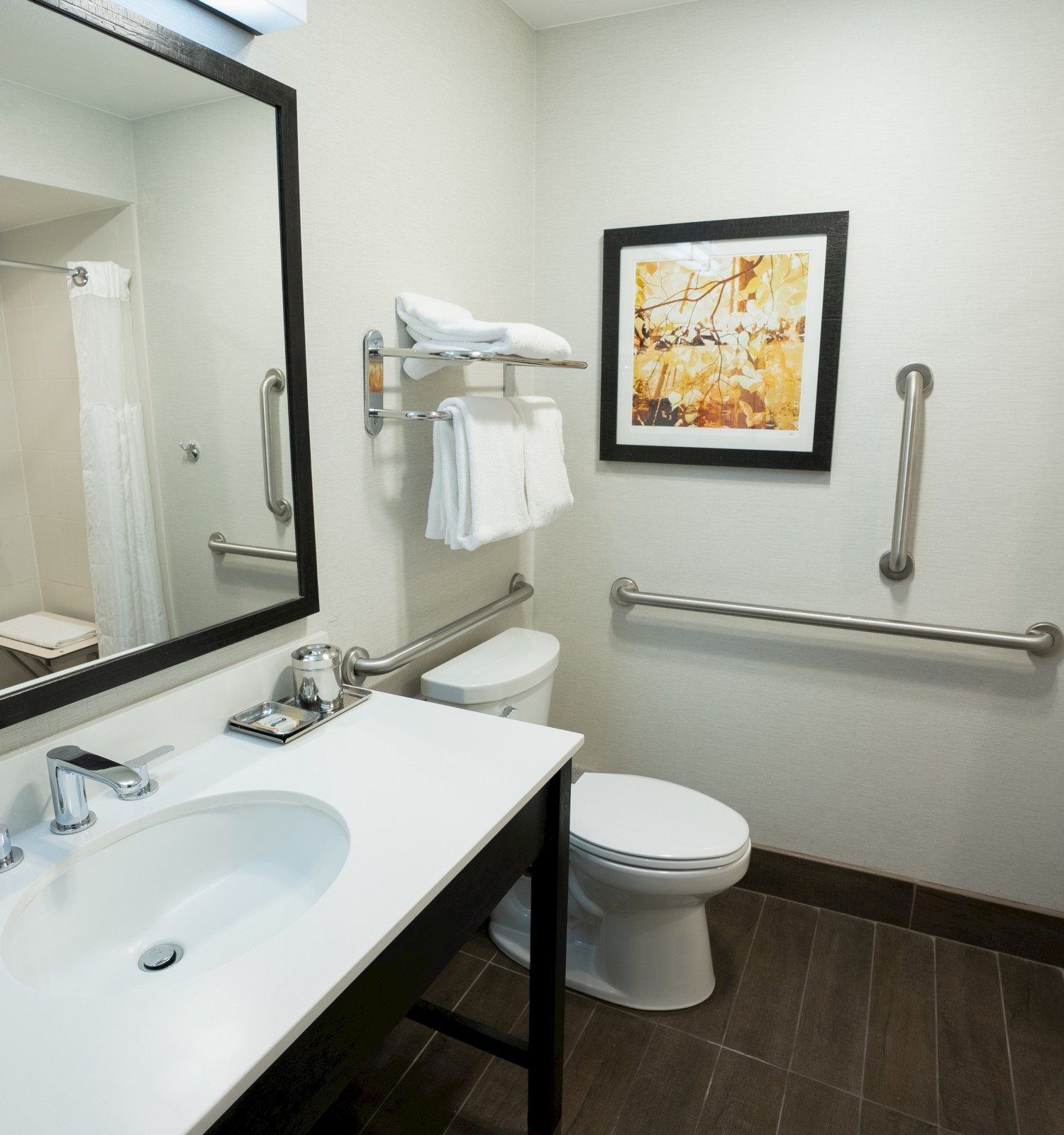 The image shows a modern bathroom with a sink, a large mirror, a toilet, a shower with a curtain, towel racks, and a framed picture on the wall.