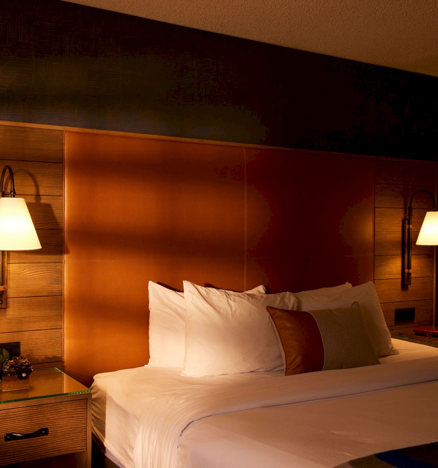 A cozy hotel room with a neatly made bed, twin bedside tables, and warm lighting from two wall-mounted lamps on either side of the bed.