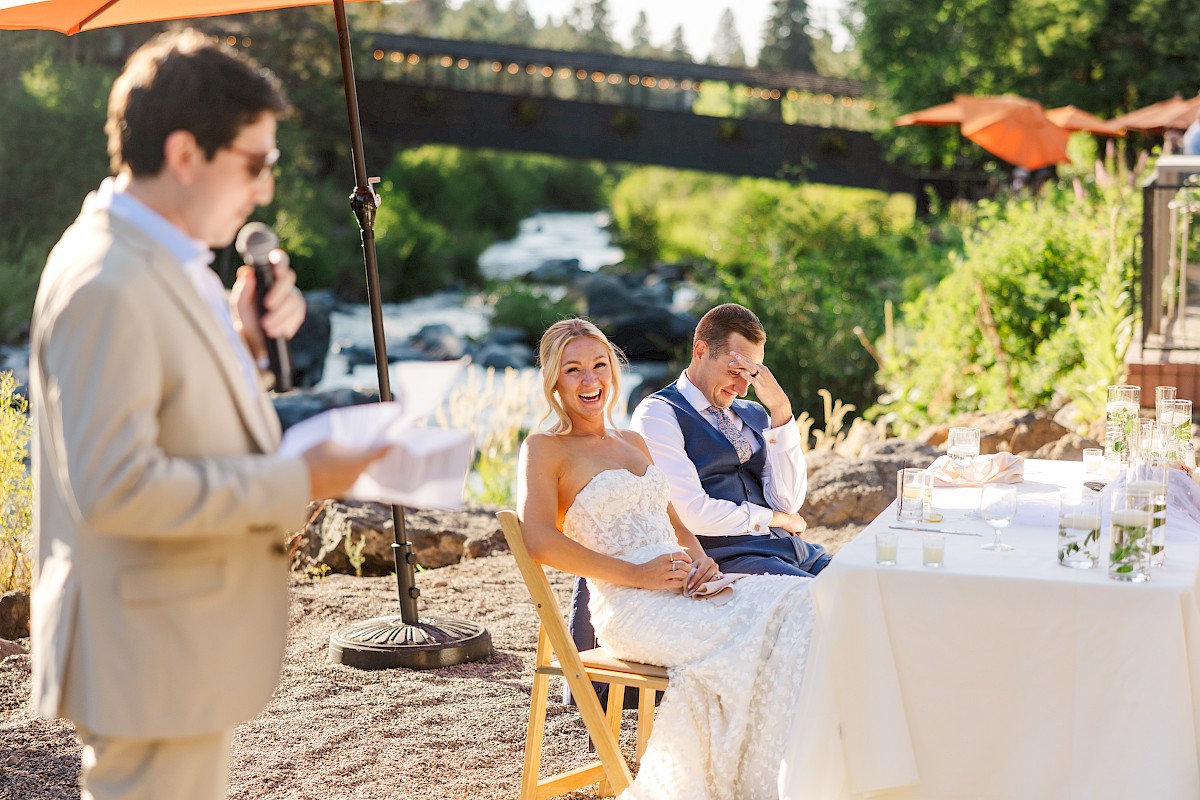 A wedding couple sits outdoors, smiling and laughing, as a man in a beige suit speaks at their reception beside a river.