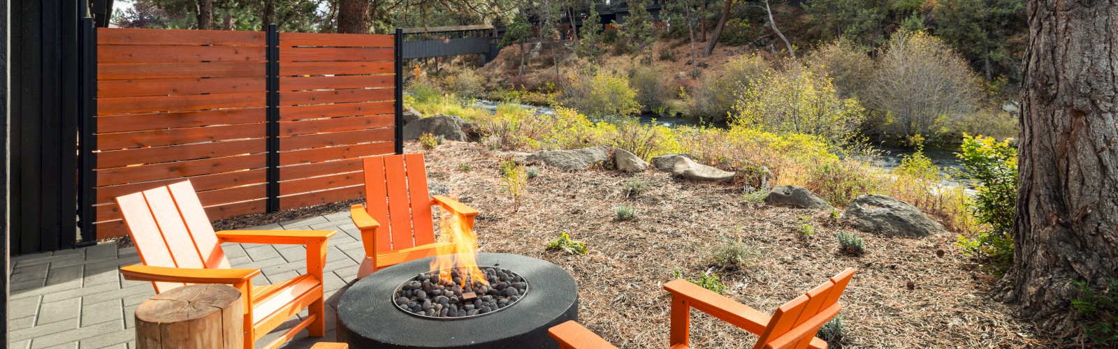 An outdoor patio features a fire pit surrounded by bright orange chairs and wooden stools, adjacent to a scenic natural landscape.