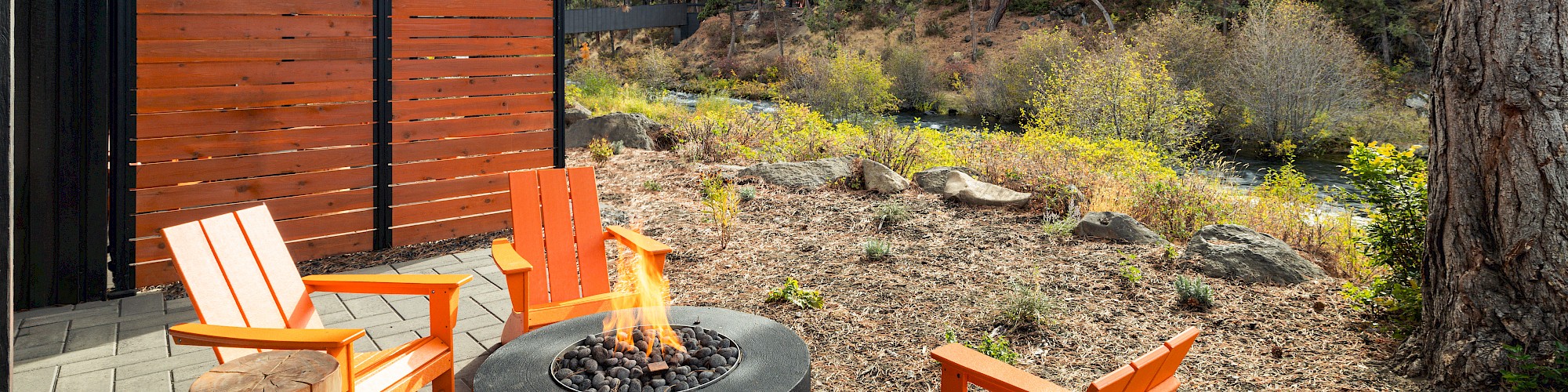 An outdoor patio features a fire pit surrounded by bright orange chairs and wooden stools, adjacent to a scenic natural landscape.