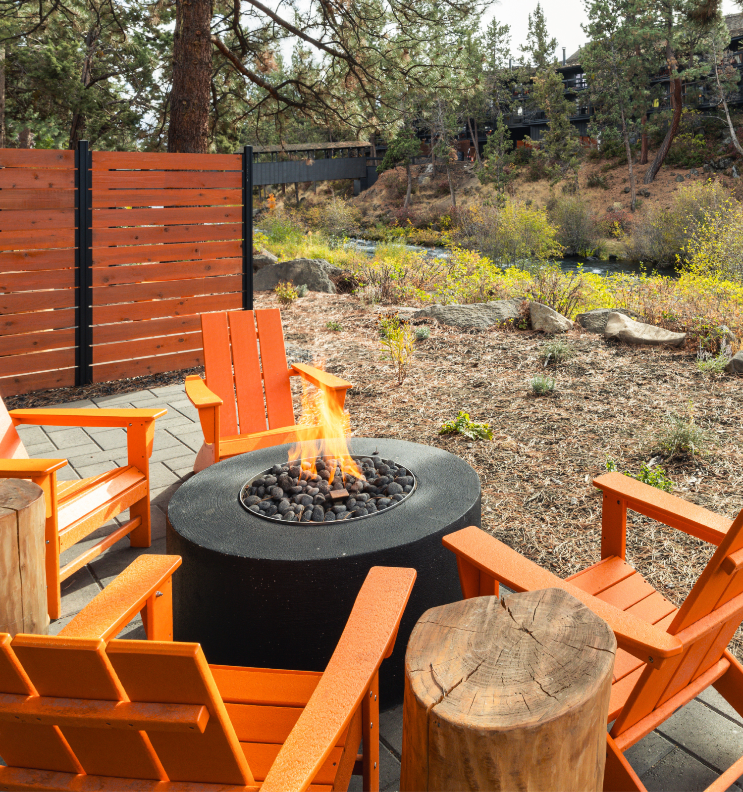 The image shows orange chairs around a lit fire pit on a patio, surrounded by trees and a wooden fence, beside a small stream.