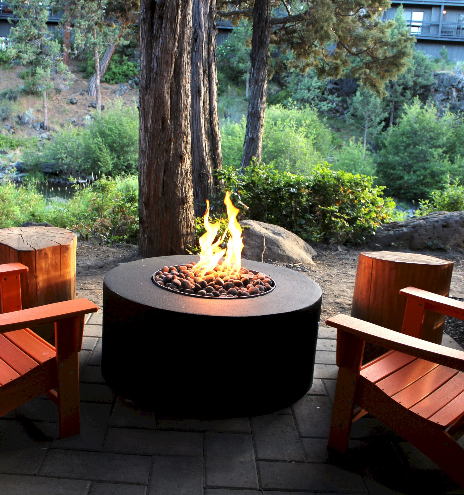 A cozy outdoor seating area features two wooden chairs and a fire pit, surrounded by trees and greenery, with a fence on one side.