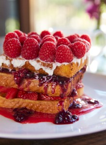 The image shows a stack of French toast with whipped cream, raspberries, and berry sauce on a white plate, served with a glass of orange juice.