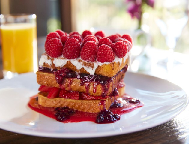 The image shows a stack of French toast with whipped cream, raspberries, and berry sauce on a white plate, served with a glass of orange juice.