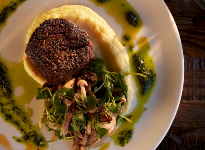 The image shows a plated dish with a piece of meat on mashed potatoes, topped with greens, and garnished with a green sauce.