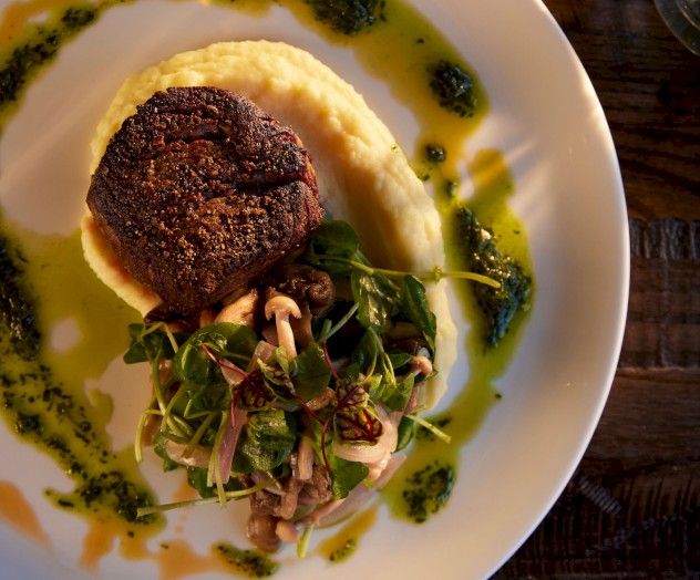 The image shows a plated dish with a piece of meat on mashed potatoes, topped with greens, and garnished with a green sauce.