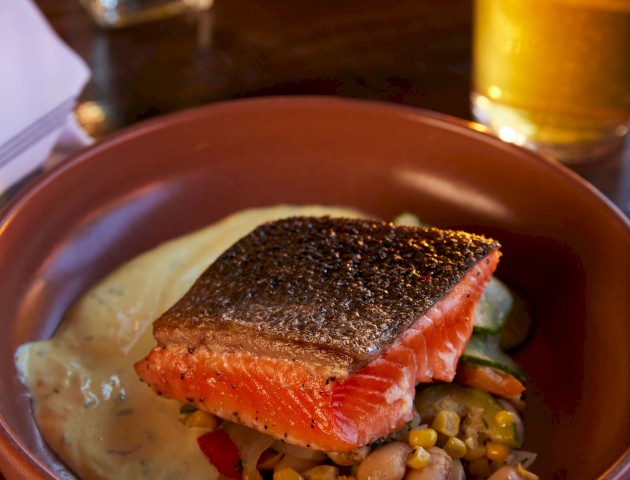 A dish with a piece of salmon, a side of mashed potatoes, and mixed vegetables in a brown bowl, accompanied by a glass of beer and another plate in the background.