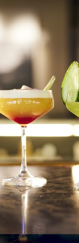 Five beverages in different glasses are lined up, including a beer, a flute with bubbly, and three cocktails with distinct colors and garnishes.