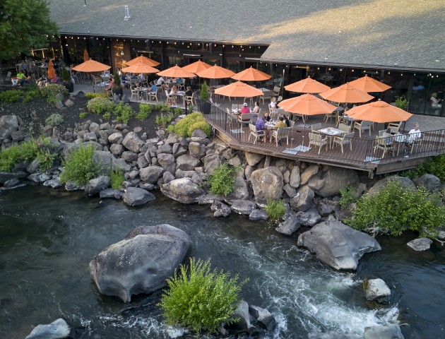 A riverside restaurant with orange umbrellas over outdoor seating is surrounded by trees and large rocks by the water, creating a serene atmosphere.