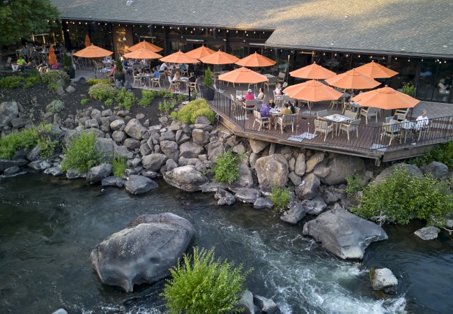 A riverside restaurant with orange umbrellas over outdoor seating is surrounded by trees and large rocks by the water, creating a serene atmosphere.