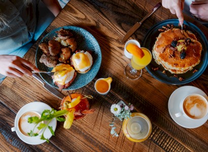 A table with brunch items: pancakes, eggs benedict, cocktails, coffee, and a small flower arrangement on a wooden surface.