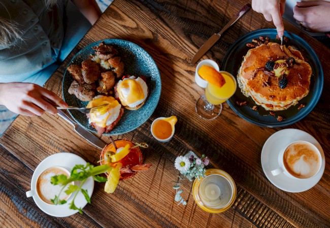 A table with brunch items: pancakes, eggs benedict, cocktails, coffee, and a small flower arrangement on a wooden surface.