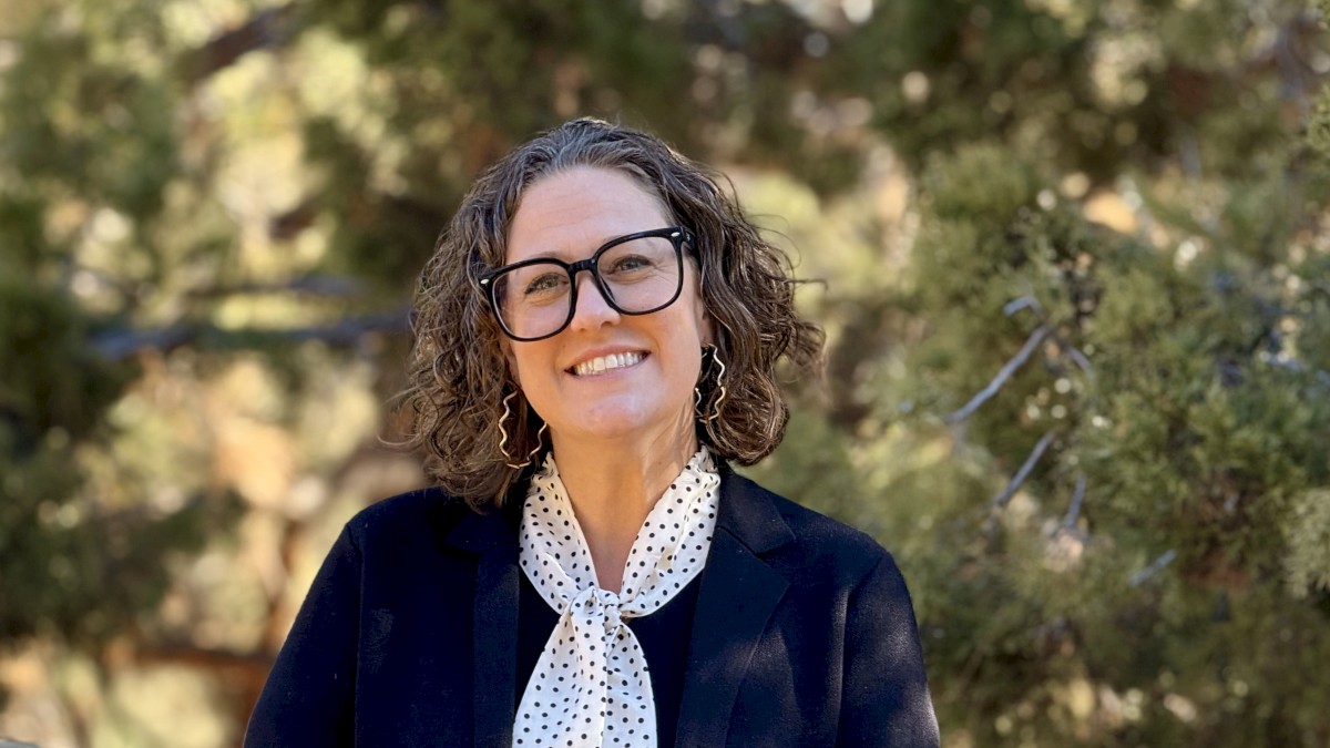 Shane Wellman Wedding and Sales Manager for Riverhouse Lodge with curly hair and glasses smiles outdoors, wearing a dark blazer and a white polka-dot scarf, standing by a wooden railing with trees behind her.