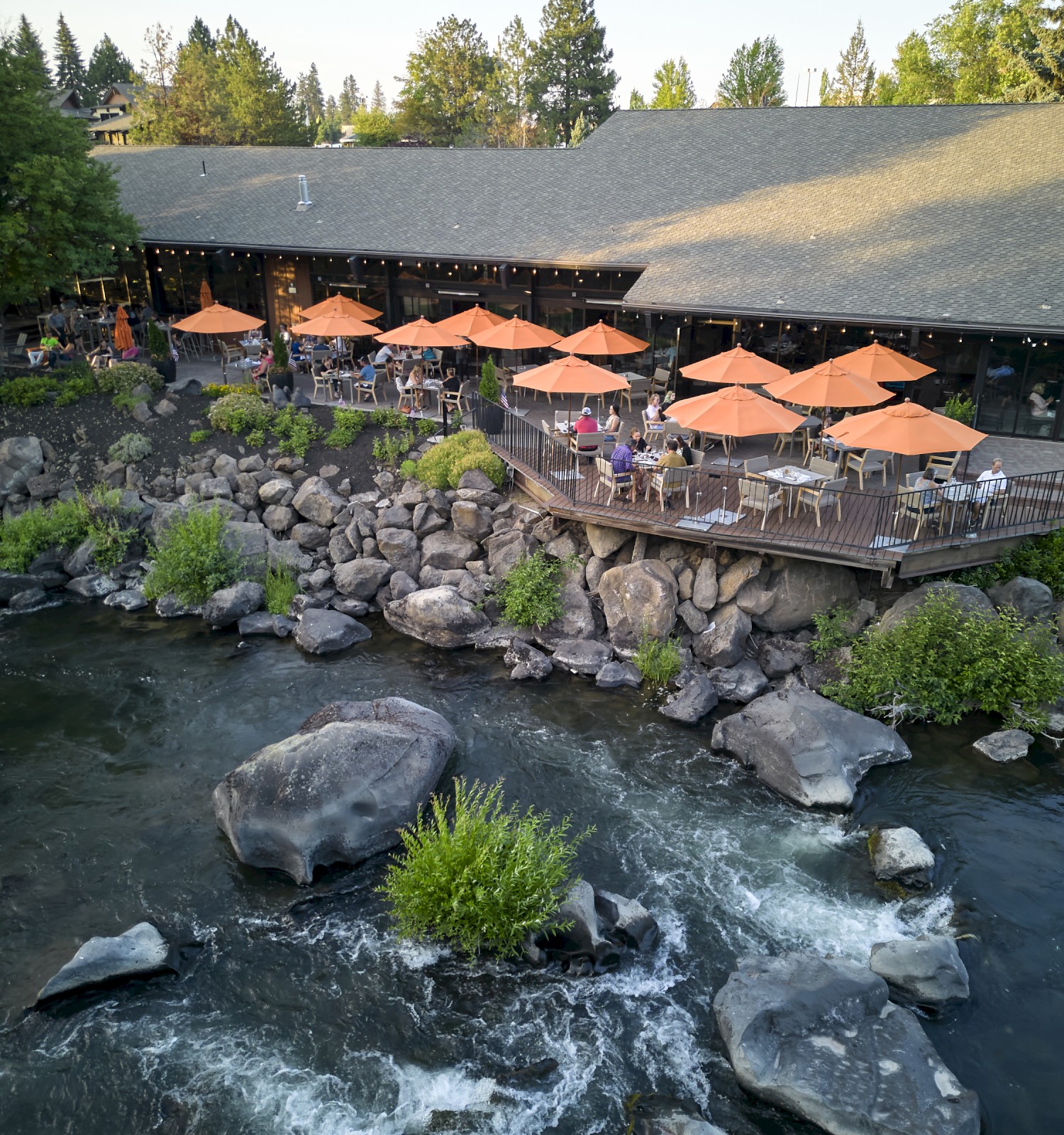 A riverside restaurant with outdoor seating under orange umbrellas, surrounded by trees and rocks, with a serene flowing river below.