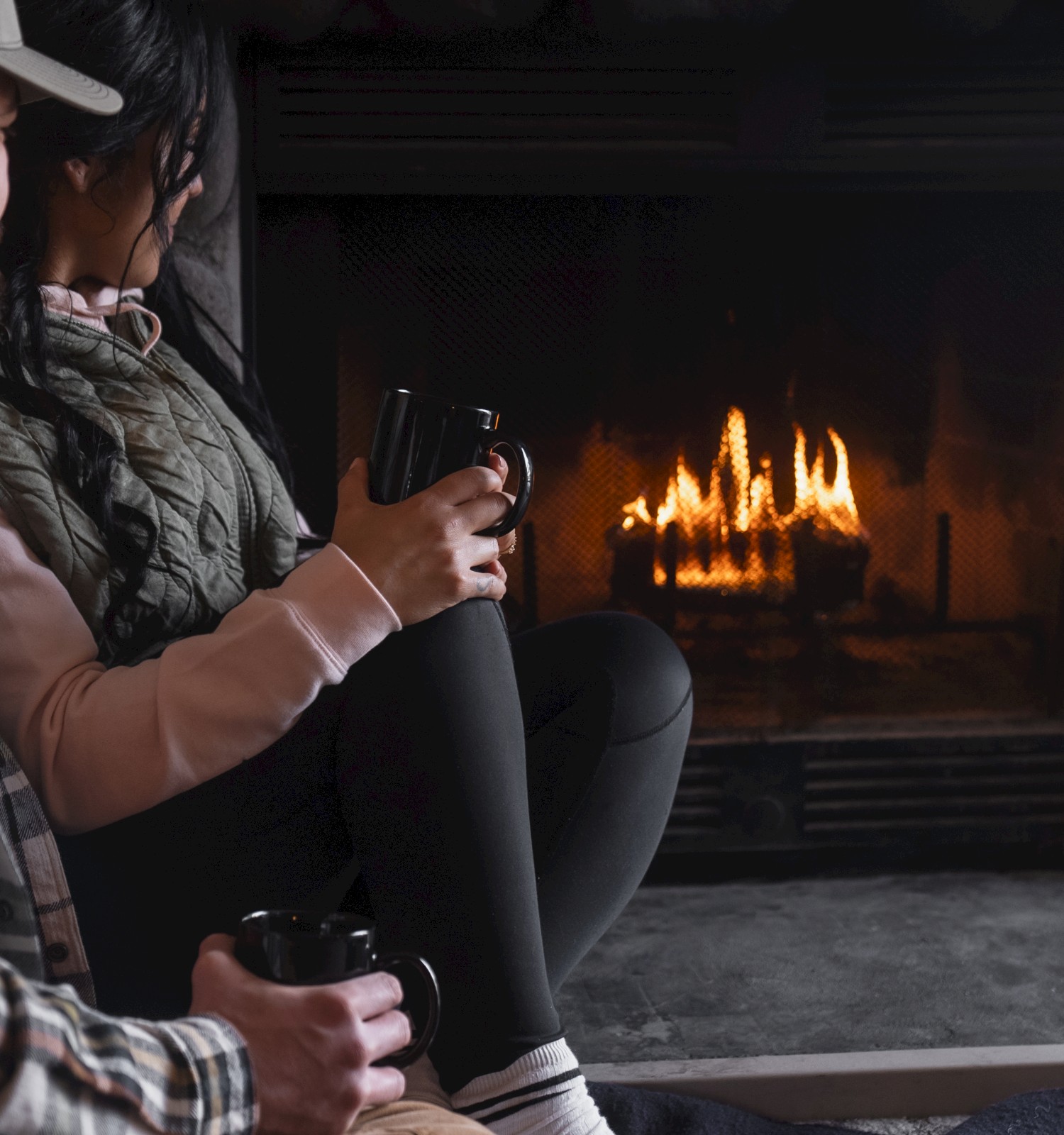 Two people sit by a lit fireplace, holding mugs and a rolled-up poster or blanket, cozy indoor scene with warm flames.