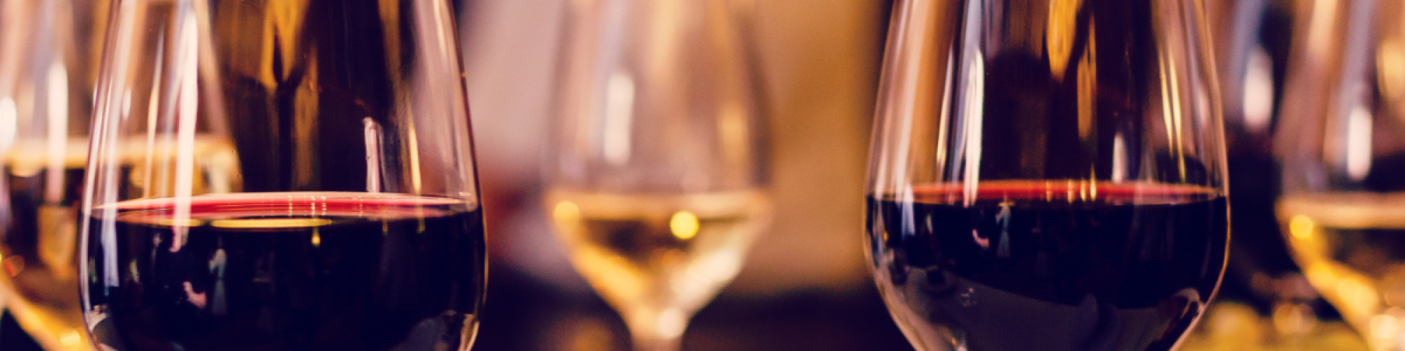 The image shows several glasses of red and white wine on a table, with a blurred background of people and warm lighting.