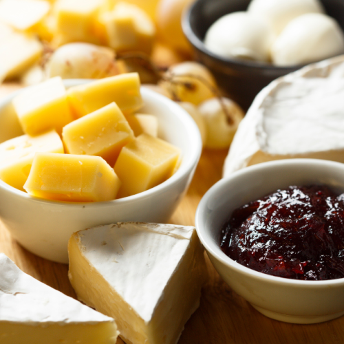 The image shows a cheese platter with various types of cheese, a bowl of jam, grapes, and raisins. There are also sliced cheese cubes in a bowl.